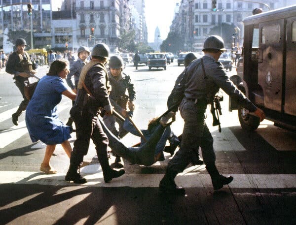Uniformed individuals carry an individual across a city street as a person in a blue dress follows closely behind. Uniformed individuals carry an individual across a city street as a person in a blue dress follows closely behind.