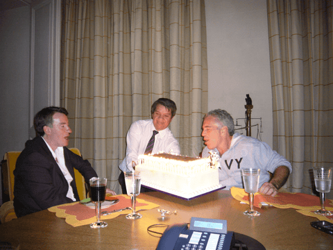 two men by a dining table as one blows out candles on a cake