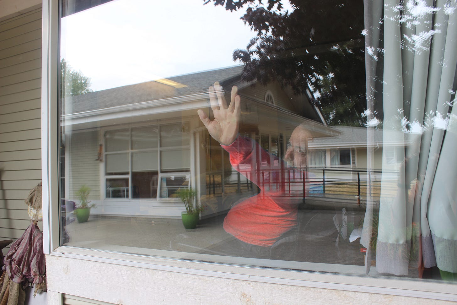 A man in a red sweater is visible behind a window pane, partially obscured by reflections. His arm is extended, and his hand is pressed against the glass in a "Live Long and Prosper" salute.