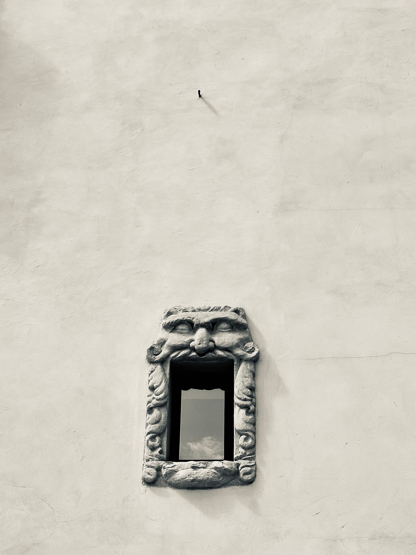 A side wall of a house has a single small window, and framing the window is a carved stone frame that resembles a gargoyle or monster into which the window becomes the inside of its mouth.