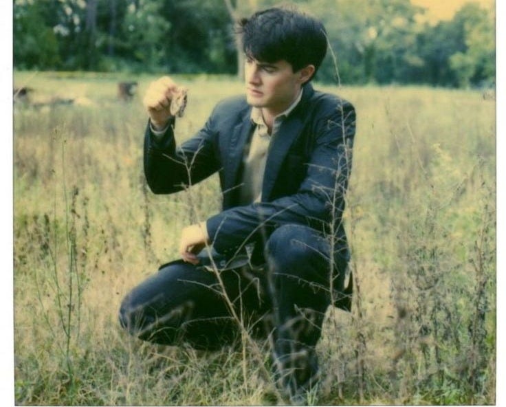This may contain: a young man sitting in the middle of a field with his hand up to his face