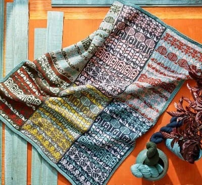 An image of a colowork blanket on an orange and turquoise background with balls of yarn to the side