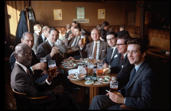 Une dizaine de jeunes gens sourient en regardant l'objectif et lèvent leur verre de bière. Nous sommes dans un. bar, autour d'une table, sans doute pour un verre entre collègues après le travail. Au premier plan, toujours le même homme noir, Omar Victor Diop, sourit, l'air un peu timide.