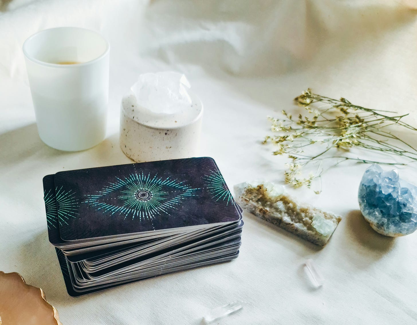 A serene flat lay featuring a stack of oracle or tarot cards with a glowing blue and green starburst pattern on the back, surrounded by a white candle, a piece of raw quartz, dried flowers, and various healing crystals on a soft cream-colored surface.