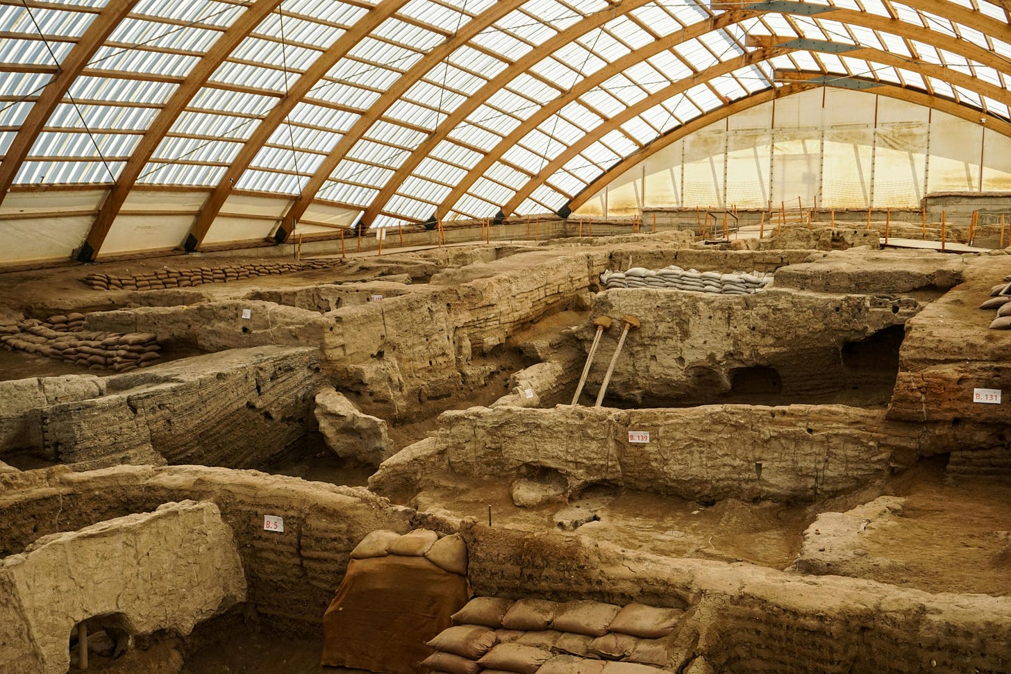 Wide interior view of the ancient archaeological ruins of Çatalhöyük in Türkiye, showing excavated mudbrick structures and preserved excavation areas beneath a large protective roof.