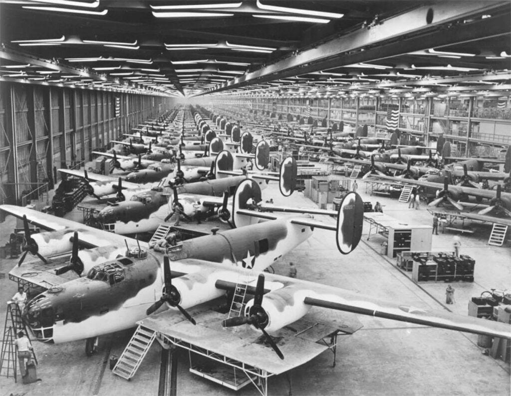 Black and white photograph of a massive aircraft manufacturing facility, showing rows of B-24 Liberator bombers in various stages of assembly. Workers can be seen as small figures among the planes, with ladders, work platforms, and equipment scattered throughout the floor.