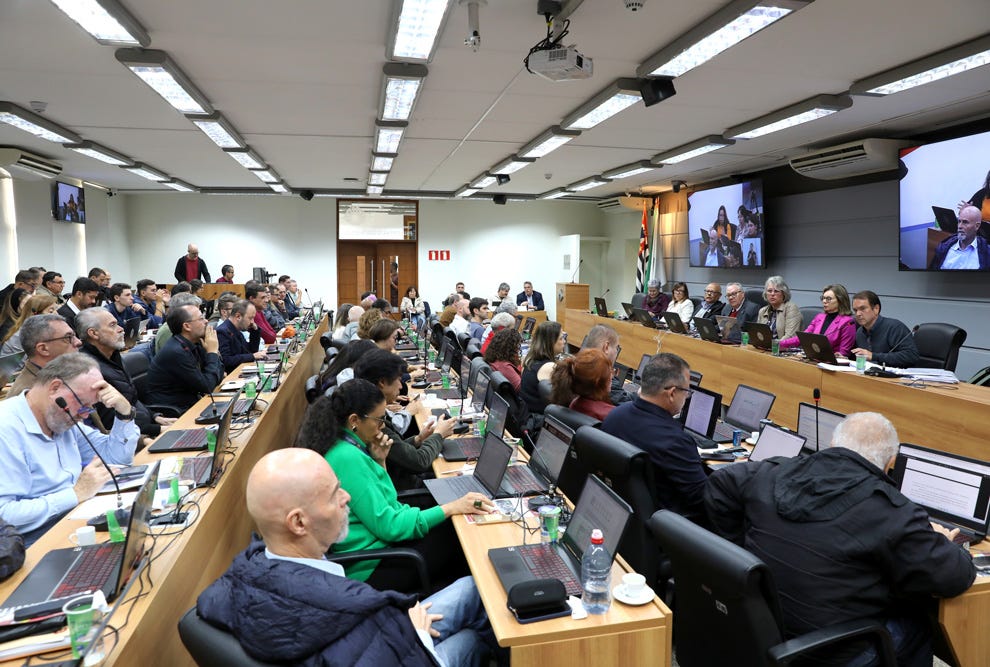 Sala de reuniões ou plenário com diversas pessoas sentadas em bancadas equipadas com laptops e microfones, enquanto uma mesa diretora ao fundo conduz os trabalhos, com telões exibindo participantes remotos e iluminação fluorescente no teto.