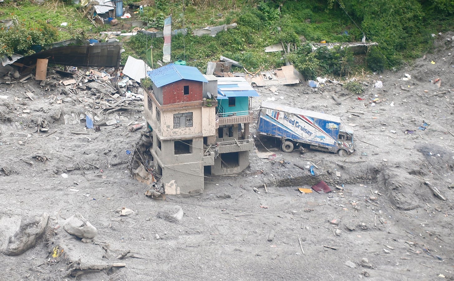 Aerial view of catastrophic landslide damage along the Nepal-Tibet border, showing a partially buried multi-story building and a stranded truck surrounded by thick mud and debris.