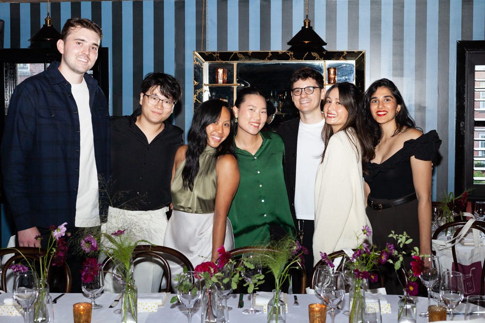 A group of seven people smiling and posing together at a dinner event. They are standing behind a long, elegantly set table with floral arrangements, candles, and glassware. The background features vertical dark-striped wallpaper and a large mirror. The atmosphere appears warm and celebratory.