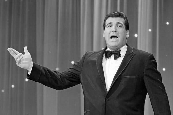 A man in a tuxedo gestures with his right hand.