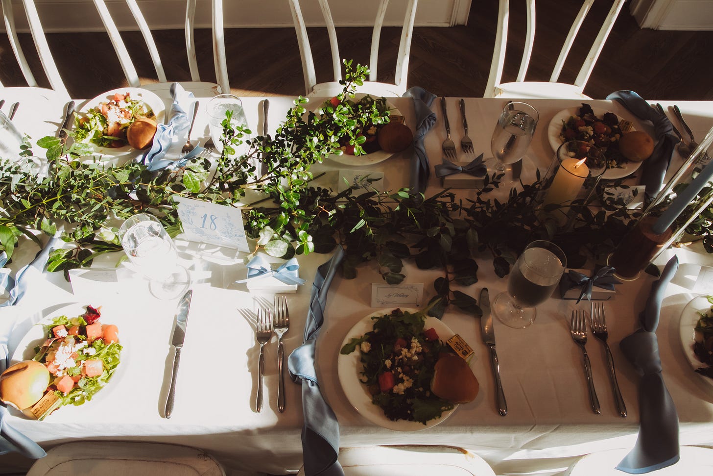 a wedding table with greenery, candles, blue napkins, and salads at every spot with watermelon on a bed of greens a wedding table with greenery, candles, blue napkins, and salads at every spot with watermelon on a bed of greens
