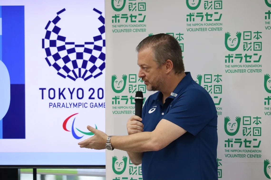 IPC President Andrew Parsons speaking into a microphone in front of a Tokyo 2020 Paralympic Games backdrop at the Nippon Foundation Volunteer Center.