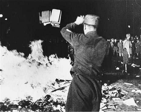 A black and white, grainy photo of a pile of books in the street, on fire. A crowd is visible looking on, and in the foreground, a Nazi soldier facing away from the camera throws several more books onto the pile.