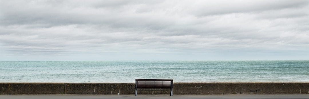 brown wooden bench on gray concrete floor near sea under white clouds during daytime brown wooden bench on gray concrete floor near sea under white clouds during daytime