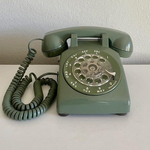May include: A vintage olive green rotary dial telephone. The phone has a circular dial with numbers and letters, a handset, and a coiled cord. The phone is set on a white surface. May include: A vintage olive green rotary dial telephone. The phone has a circular dial with numbers and letters, a handset, and a coiled cord. The phone is set on a white surface.