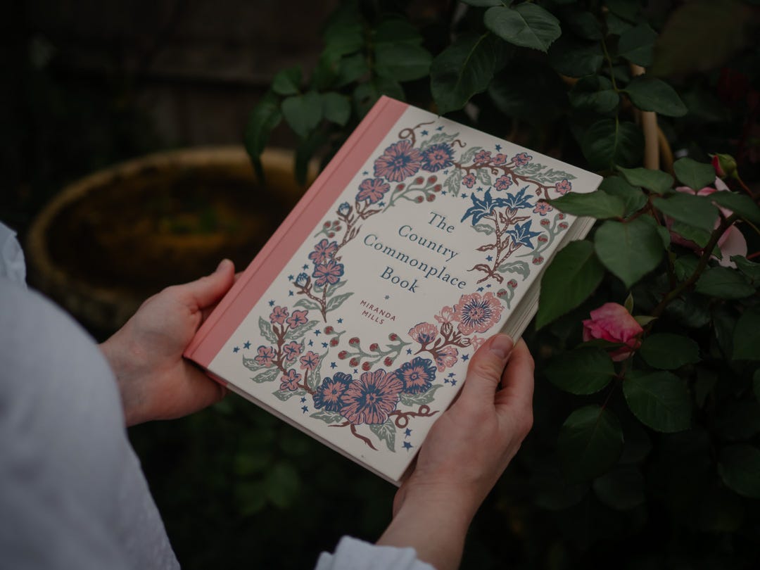 Female hands holding The Country Commonplace Book by Miranda Mills in a garden with roses behind.