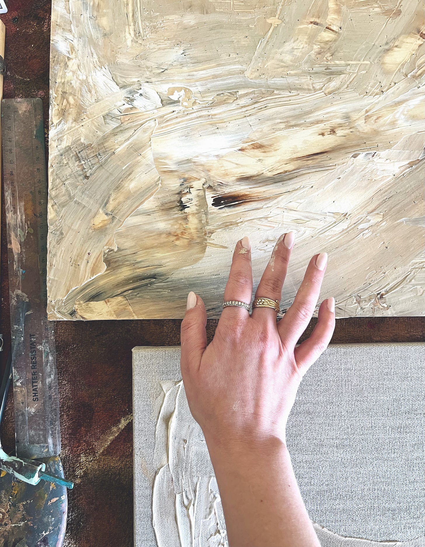 A close-up of the artist’s hand resting on a softly textured, abstract painting in earthy tones. The gesture is quiet and grounded, with soft natural light falling across the canvas.