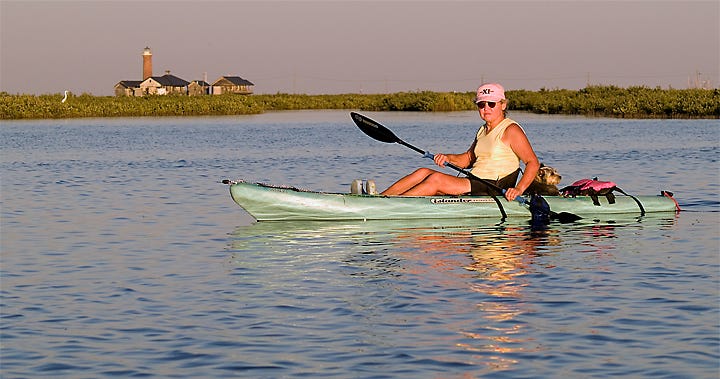 Photo of a woman and a dog in a kayak on the water, with a lighthouse and a great white heron in the background.