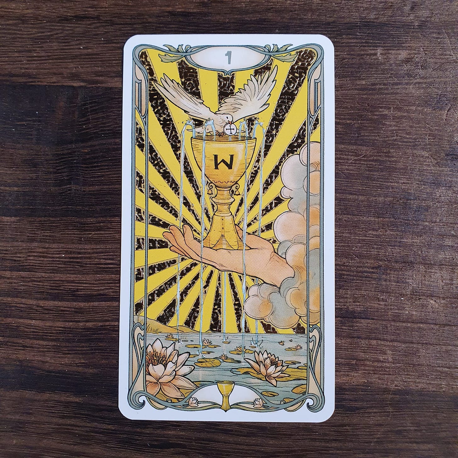 Ace of Cups from the Golden Art Nouveau Tarot showing chalice overflowing with water and descending dove, placed on wooden table for the Tarot DMs interview with Alice Slater by Finbarre Snarey.