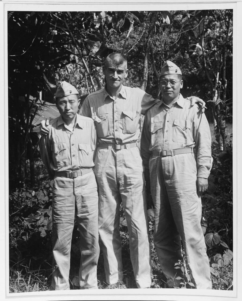 Outdoor black-and-white photograph of LtCol Evans F. Carlson standing in jungle terrain with two interpreters, arms around their shoulders, all wearing light-colored field uniforms during a World War II Pacific campaign.