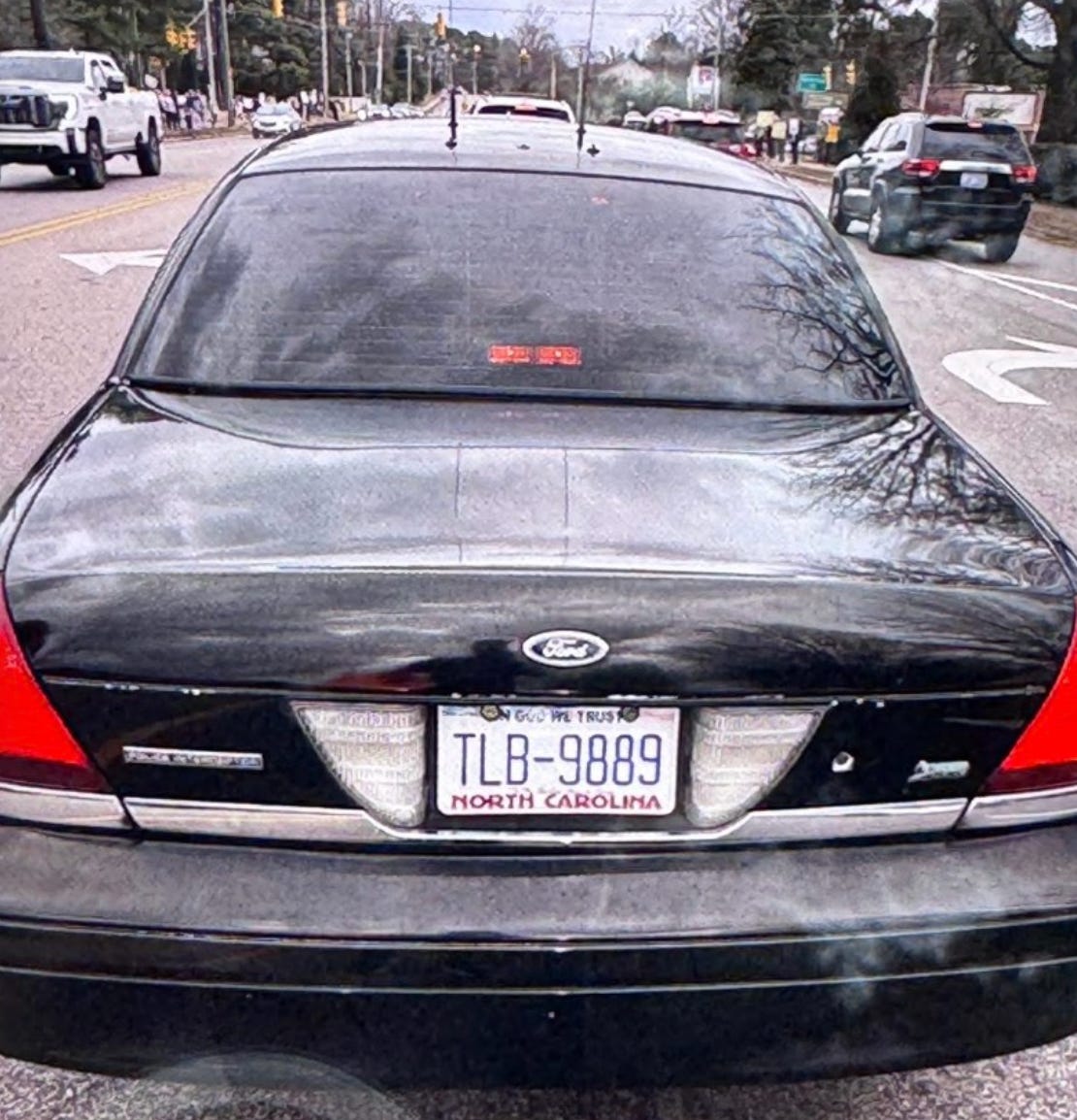 A rear view of a black Ford Crown Victoria Police Interceptor driving on a paved road. The car has dark tinted windows, dual antennas on the trunk, and a North Carolina license plate reading "TLB-9889" with "In God We Trust" at the top. The "Police Interceptor" badge is visible on the left side of the trunk. In the background, other cars and trees are visible on a street during the day.