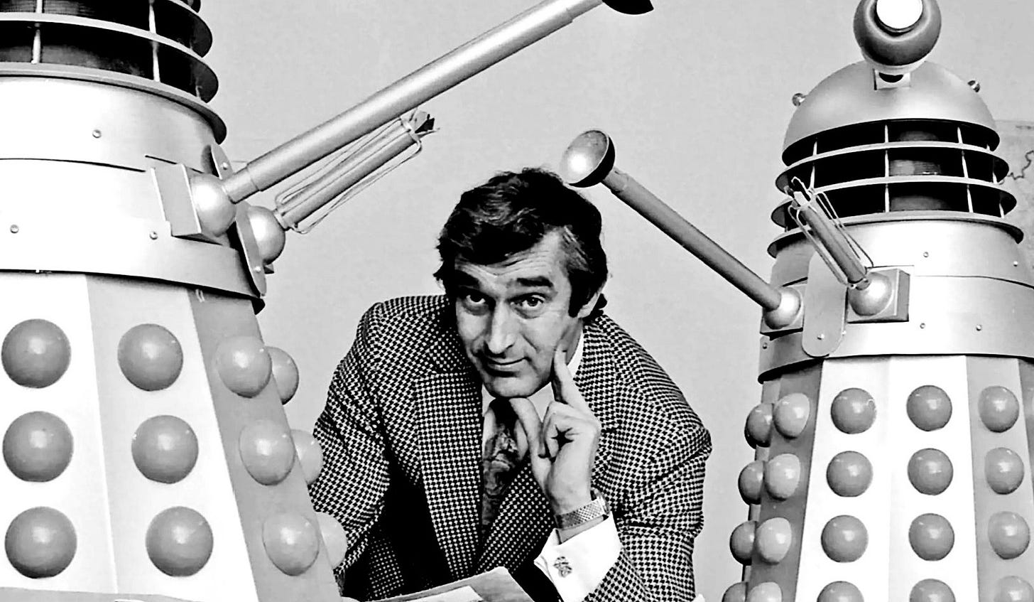 Terry Nation in early middle age wearing a natty suit and flanked by a pair of large-scale model Daleks.