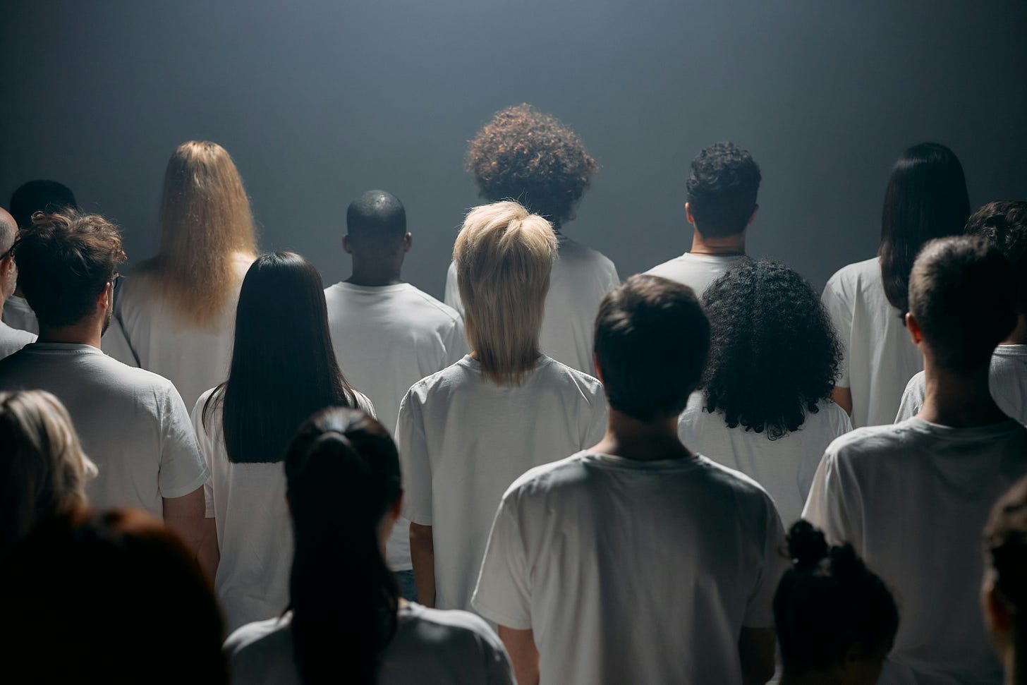 A imagem mostra um grupo de pessoas em pé, muito próximas umas das outras, todas voltadas de costas para a câmera. Elas estão vestindo camisetas brancas simples e idênticas, o que cria uma sensação de uniformidade. A iluminação vem de cima, como um foco de luz, projetando sombras marcantes e criando um clima dramático e introspectivo. O fundo é escuro e minimalista, sem elementos visuais que distraiam. Isso faz com que as pessoas sejam o centro absoluto da imagem. Apesar da roupa igual, há diversidade entre os indivíduos: os cabelos variam em cor, textura e estilo — alguns são lisos, outros cacheados, curtos ou longos — sugerindo que são pessoas diferentes, com identidades únicas. O fato de estarem de costas transmite uma sensação de anonimato, como se fossem uma massa coletiva, mas ainda composta por indivíduos distintos.