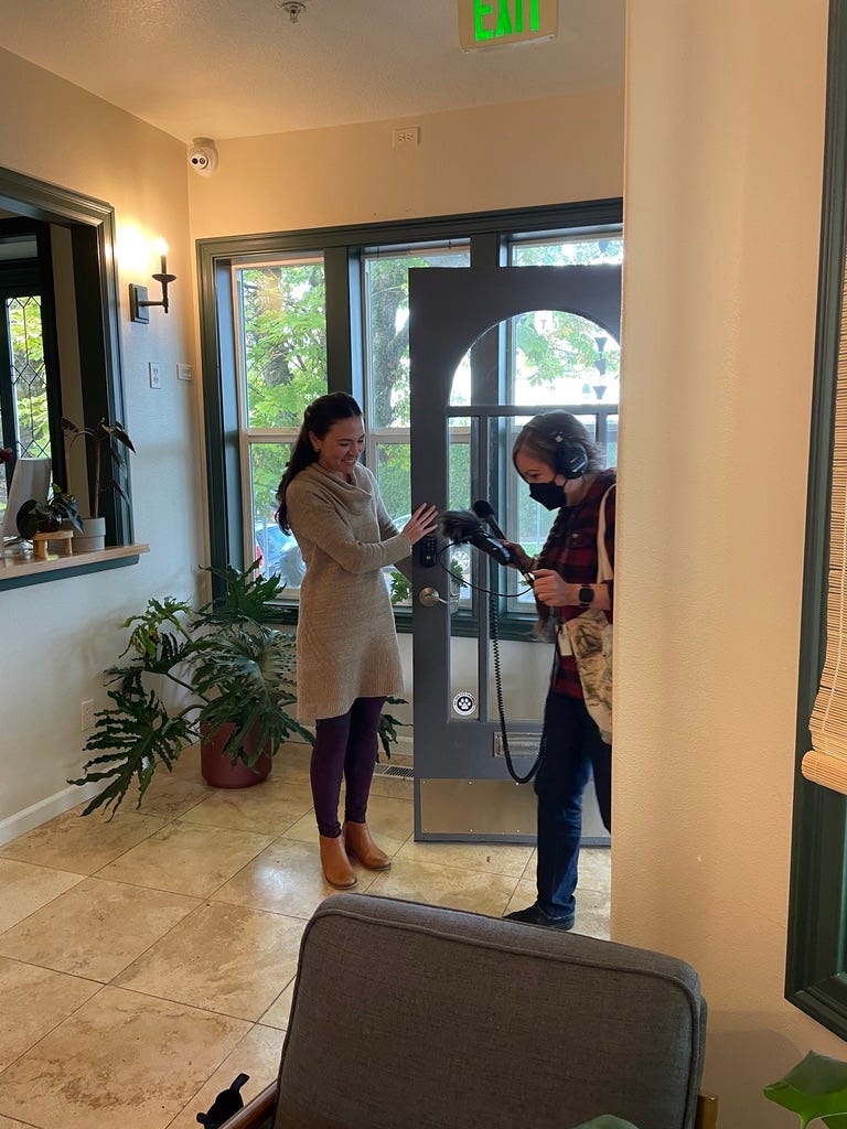 A person holding audio equipment enters a building lobby as another person holds open the door, smiling.