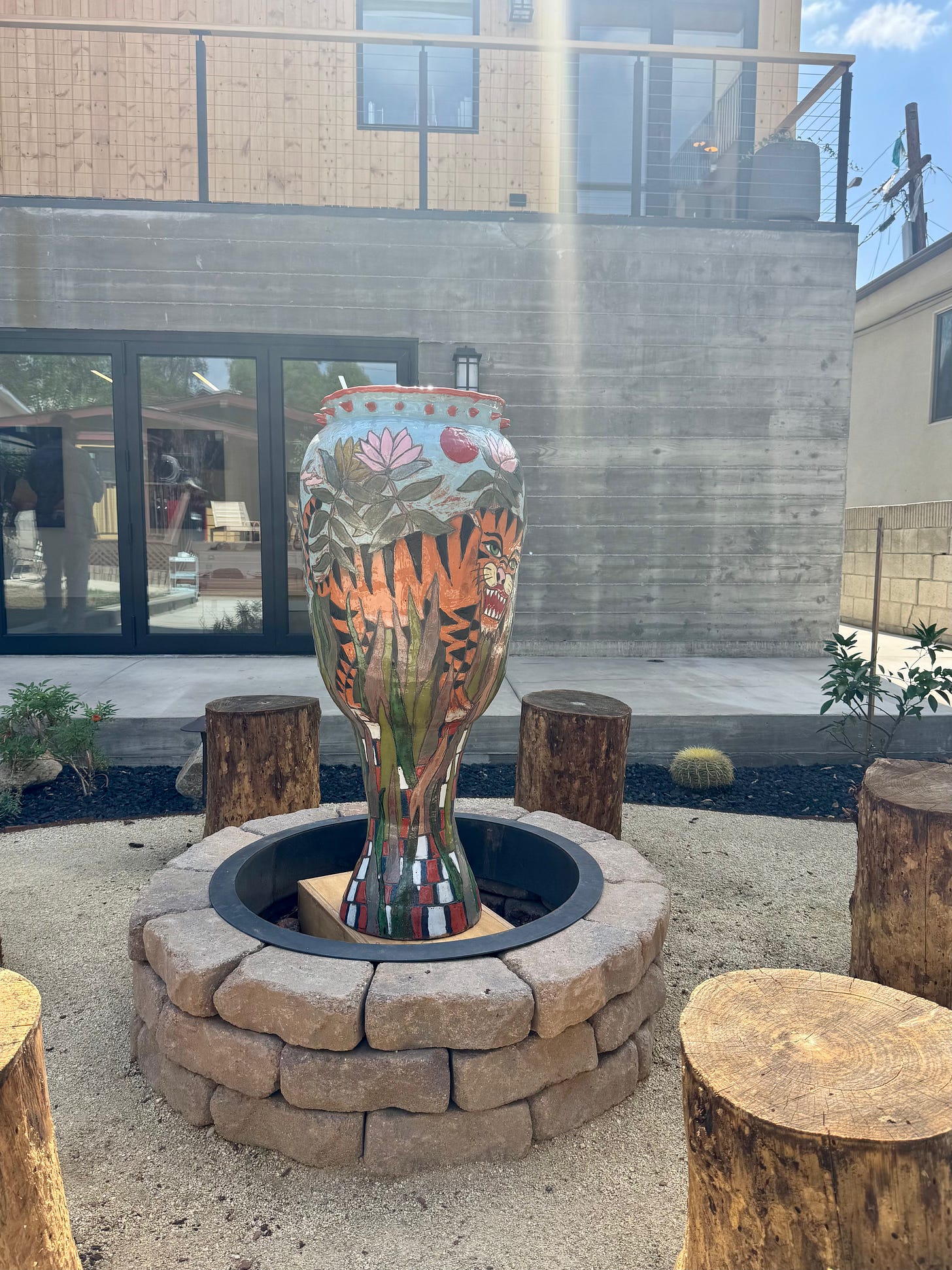 Tall painted ceramic vessel by Patrick Arnold standing in a circular stone ring in the gravel courtyard at Feia in Highland Park, with tree-stump seats and the gallery building behind.