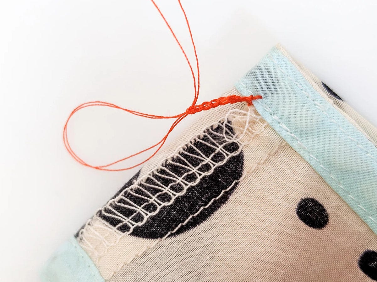 Needle pulling orange thread through fabric near armhole seam with serged edge and light blue binding.