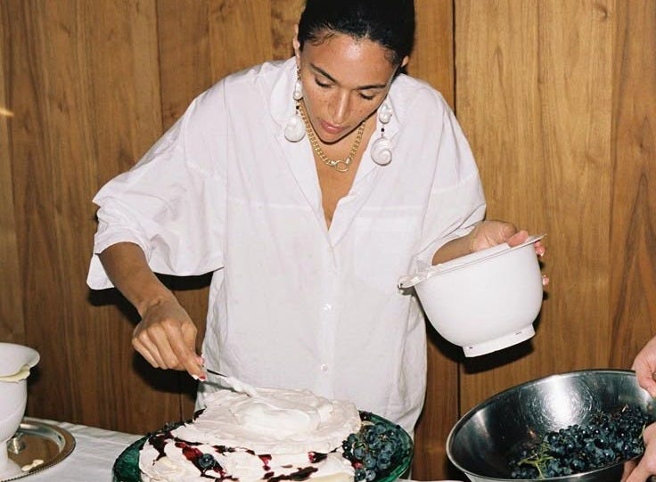 This may contain: a woman in white shirt decorating cake with icing on table next to wooden wall