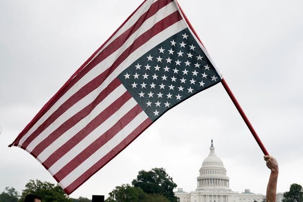 A man holds a flagpole with an upside-down American flag with the U.S. Capitol visible in the distance.
