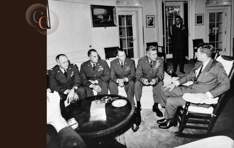 A black and white photograph of President John F. Kennedy seated in a rocking chair, facing four senior military officers on a sofa in the Oval Office during the Cuban Missile Crisis. The Career Communiqué logo appears in the upper left corner.