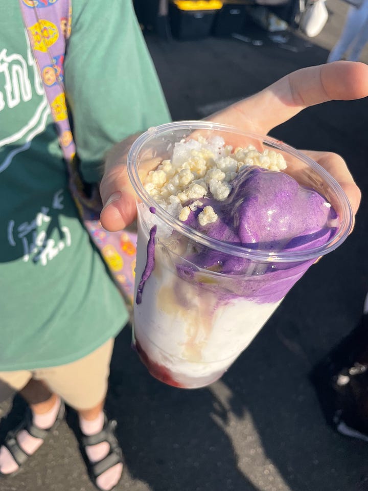man holds cup of purple ice cream and coconut cream with red beans at the bottom, a pickle and some sauerkraut in front of a food tent