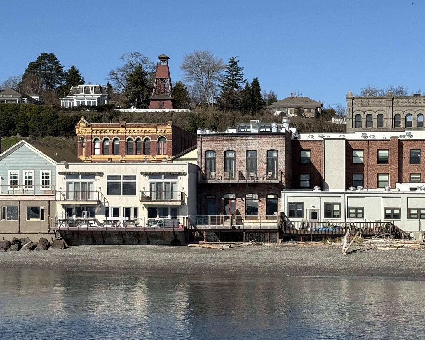 A view from across water of a 75-foot wooden fire bell tower and historic homes line the bluff above the intact 19th-centure commercial architecture of downtown, Port Townsend, Washington