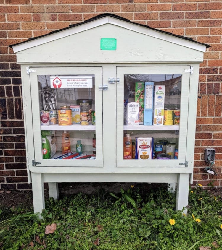 A standalone gray cabinet outside. Through panes of glass, in the cabinet there are non-perishable food items and personal care items.