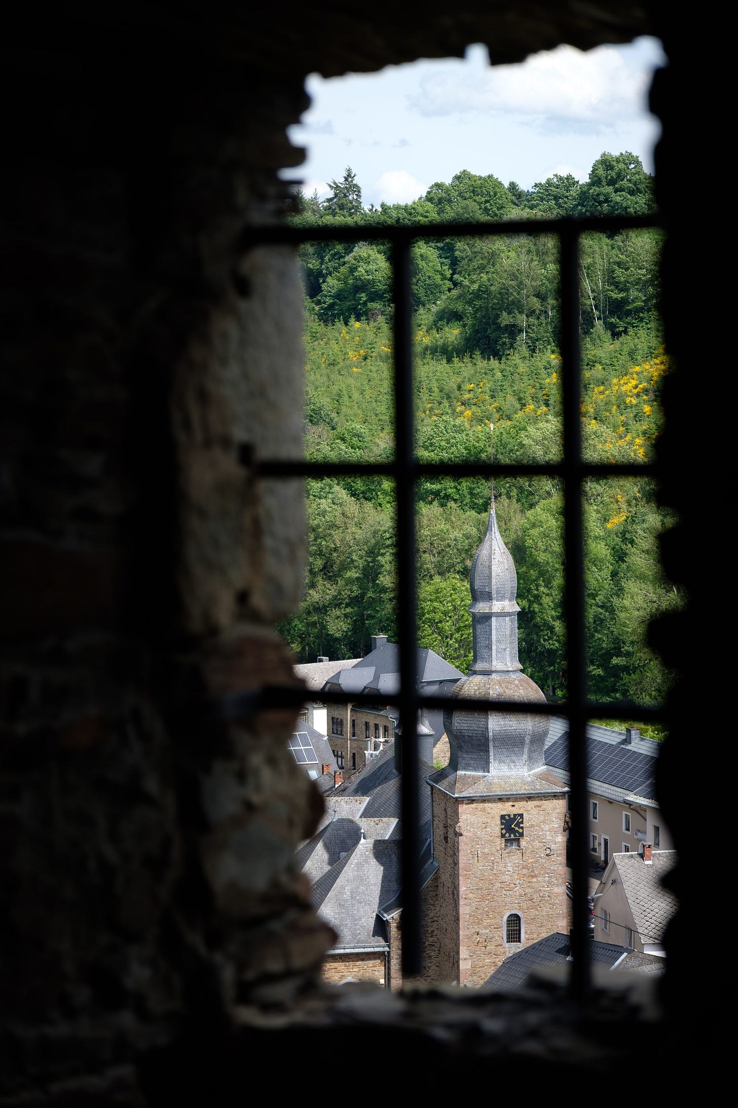 A photograph taken from inside a medieval stone tower, looking out through a narrow barred window onto an ancient town. Sunlight spills into the view, creating a soft glow that contrasts with the dark interior.