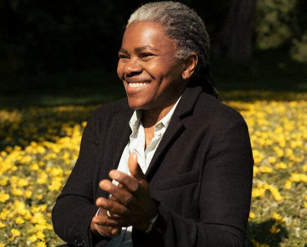 A woman with graying dreads sits in a field of yellow flowers, smiling and clapping her hands together while wearing a light button-up shirt, jeans and a dark blazer.