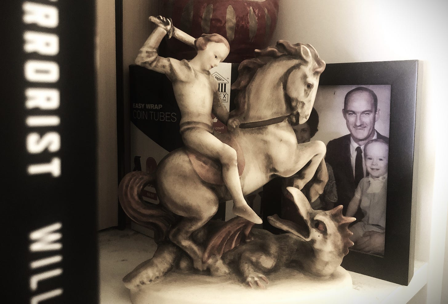 The same Hummel figurine sits on a shelf, grimy with age, partially obscuring a framed portrait photograph of a family of four. Only a small boy in the front row and his father behind are visible, dressed as if from the early 1970s. In the extreme foreground is the blurry spine of a book, which we can guess from the "RRORIST WILL" is The Accidental Terrorist by William Shunn. The same Hummel figurine sits on a shelf, grimy with age, partially obscuring a framed portrait photograph of a family of four. Only a small boy in the front row and his father behind are visible, dressed as if from the early 1970s. In the extreme foreground is the blurry spine of a book, which we can guess from the "RRORIST WILL" is The Accidental Terrorist by William Shunn.