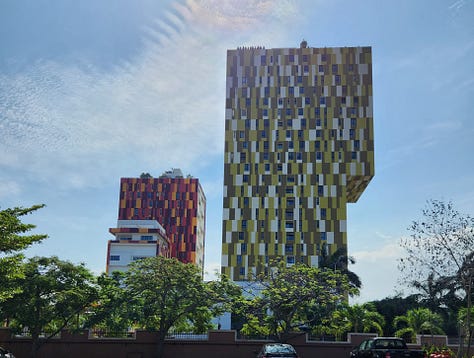 Two multicolored high rise apartment buildings, a Burger King sign near a stairwell in the mall, and a table full of fresh plantains on a roadside vendors table.