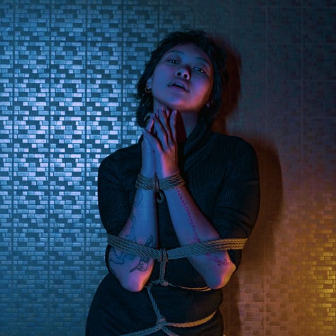 A girl dressed in black is posing with confidence, tied in ropes, over a blue and orange tiled background.