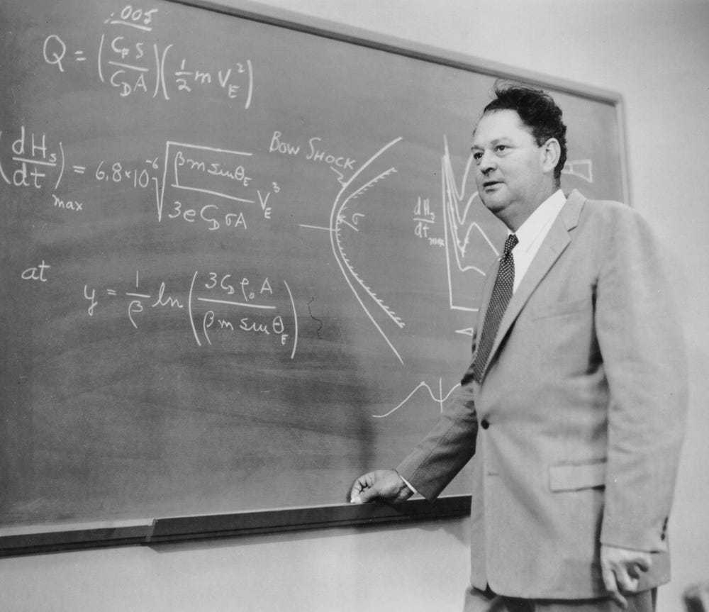 A black-and-white photograph of a man in a suit and tie standing at a chalkboard covered in aerodynamics or atmospheric re-entry equations and diagrams. The equations reference concepts such as heat transfer rate (dHs/dt), bow shock, drag coefficient (CD), and kinetic energy (½mVE²). The setting appears to be an academic or research lecture environment, likely from the 1950s or 1960s based on clothing and photographic style.