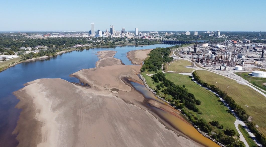 An opaque, bright orange plume encroaches on a large sandbar from the west (right) bank of the Arkansas River. Along the shoreline, a band of leafy trees separates the river from a large petroleum refinery. Downtown Tulsa is visible downstream, opposite the refinery.
