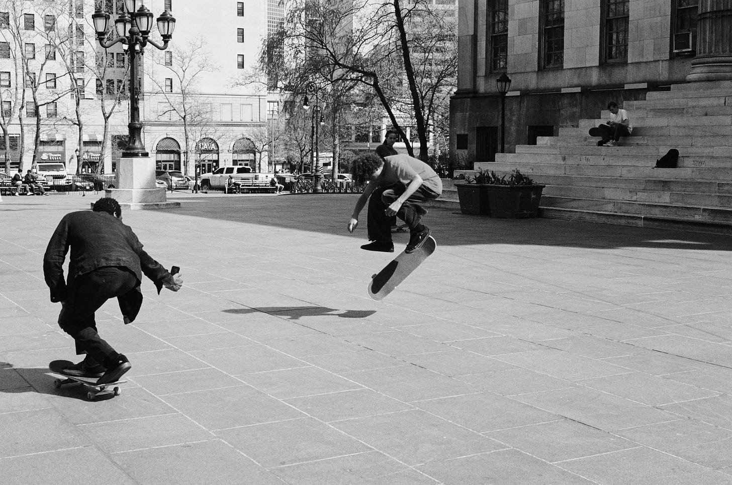 A father skateboards while filming his son mid-air doing a trick on his skateboard in a public plaza