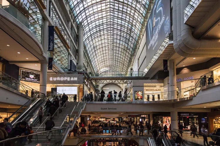 A photo of the CF Eaton Centre in Toronto. Multiple escalators and storefronts are shown, and the space is packed with consumers. Light streams in from the glass roof, and trifoliums are present on the upper levels, emphasizing the perceived grandeur of the building.