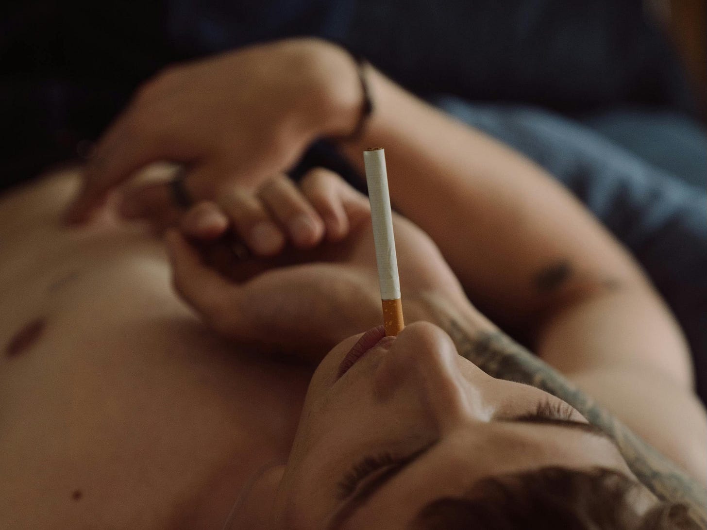 Two people in an intimate embrace, one resting on the other's chest with an unlit cigarette, fingers touching. Photo by Cottonbro Studio.