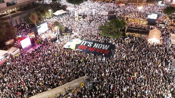 A large crowd gathered at night in an urban setting. People are densely packed, holding a prominent banner with "IT\'S NOW OR NEVER" in bold red and black text. Bright lights and screens are visible, illuminating the scene.