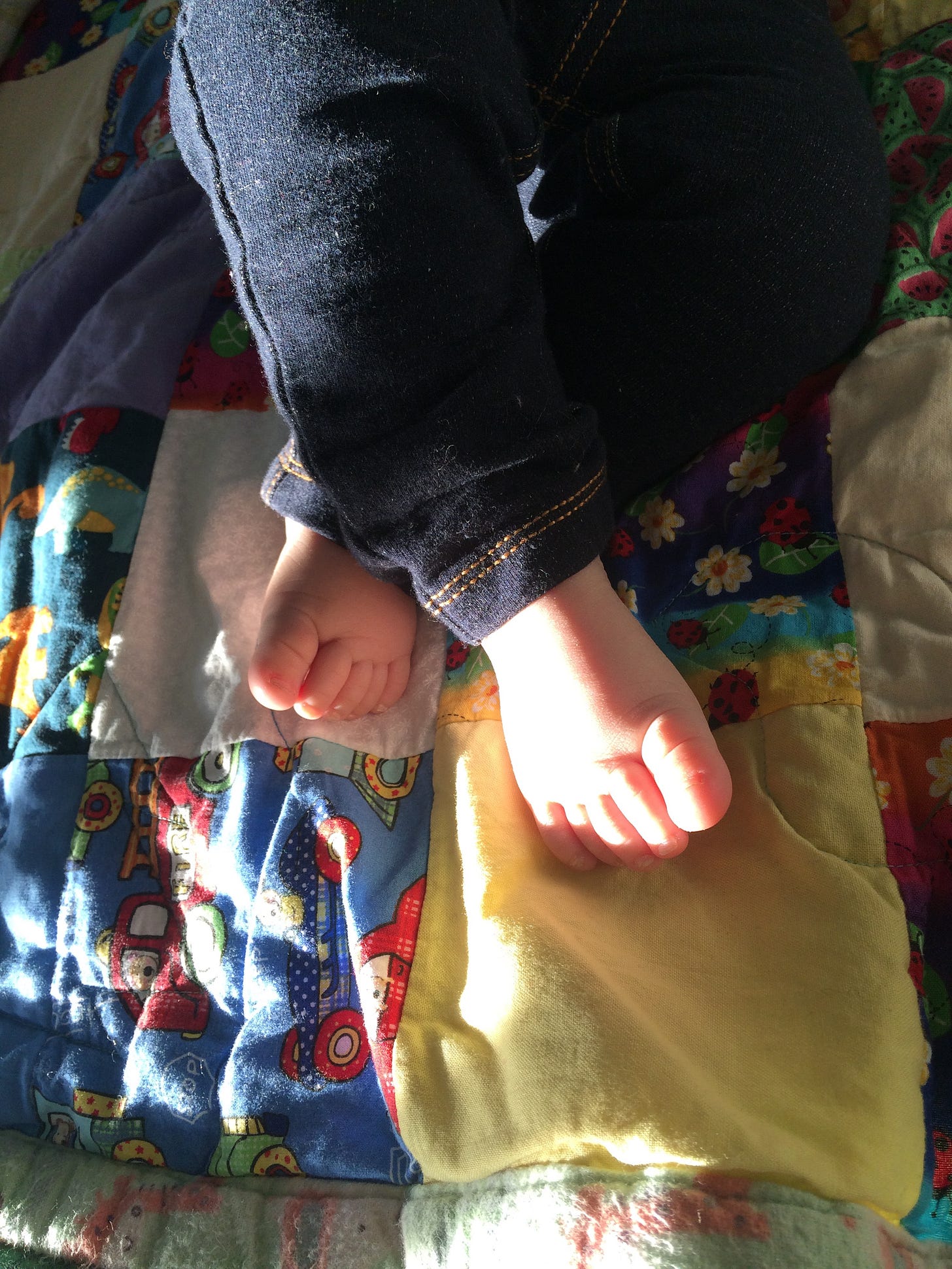 An image of baby legs in baby blue jeans, their bare feet against a rumpled quilt in a shaft of sunlight