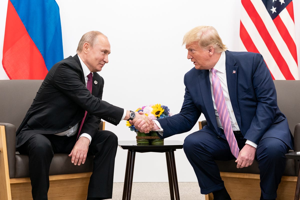 Trump and Putin, both seated, lean over and shake hands Trump and Putin, both seated, lean over and shake hands