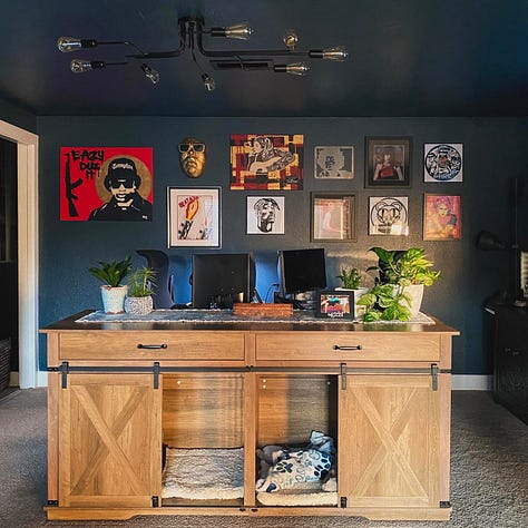 Home office with dark blue walls, light wood desk, plants, computers, and a gallery wall featuring hip-hop art like Eazy-E and Tupac Shakur, and a gold mask. Shows a maximalist, personal decor style.